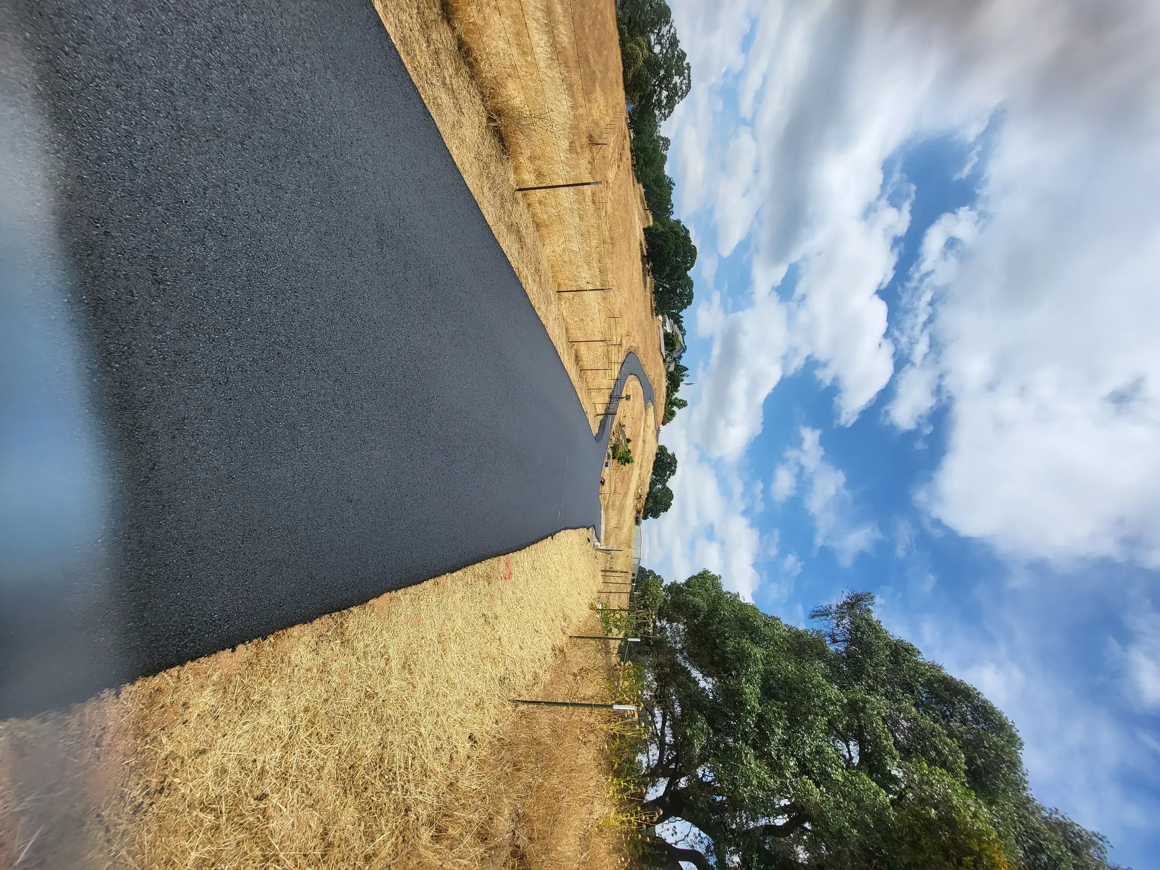 Newly paved asphalt driveway with dark seal coat and defined edges adjacent to gravel and vegetation